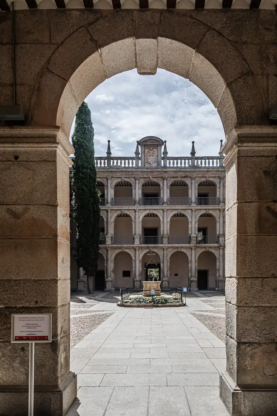 Patio de tres alturas con arcadas, fuente y ciprés visto a través de un arco de piedra, Universidad de Alcalá