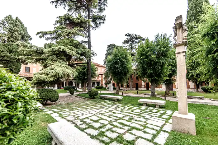 Patio ajardinado con crucero de piedra, bancos y cedros del recinto de San Ildefonso